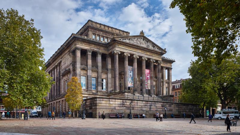 A neoclassical building with tall stone columns and ornate pediment, surrounded by trees and a paved square where people are walking.