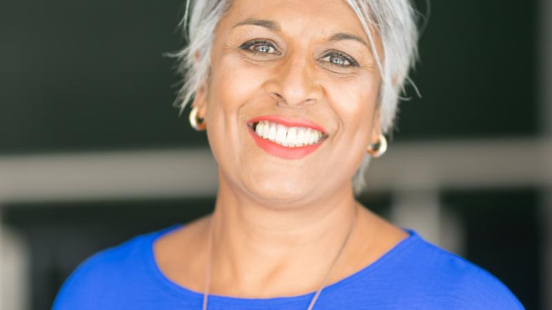 Woman with brown skin wearing a blue top, short grey hair and smiling.