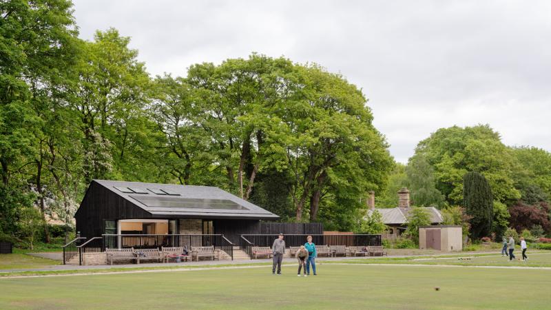 People playing crown green bowls on a green in a park setting, with a pavilion building behind.