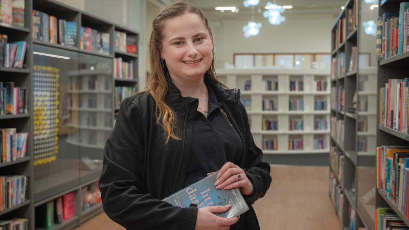 A young lady with long brown hair, wearing black and holding a book in a library and smiling. 