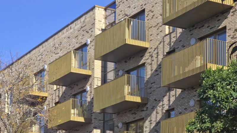 A residential apartment block over several floors with light brick and gold balconies, framed by trees.