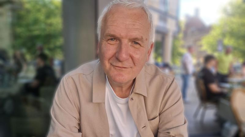 A man with grey hair, wearing a beige open first and white t-shirt underneath sat outside smiling at the camera