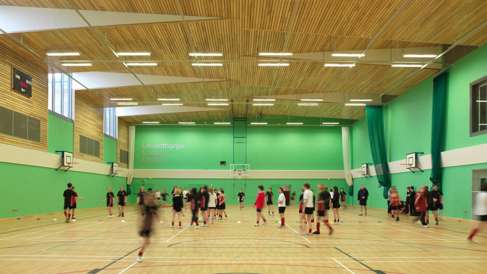 a sports hall with green walls and timber roof full of children playing sport