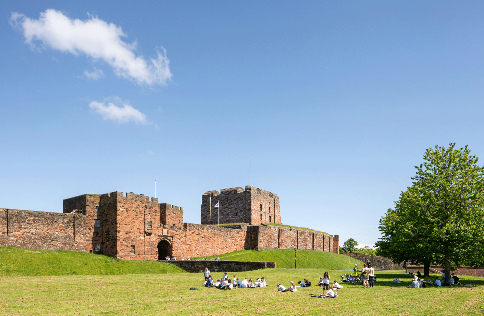 A group of schoolchildren sit on the grass in front of Carlisle Castle on a sunny day