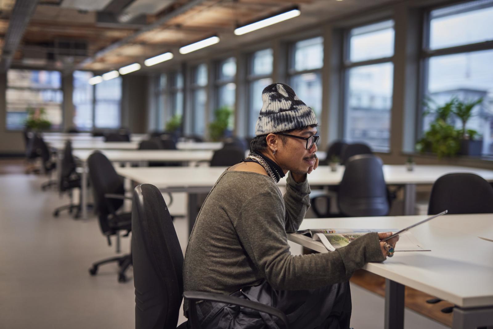 a man wearing a beanie sits at a table with an ipad in an office at Hilton House.