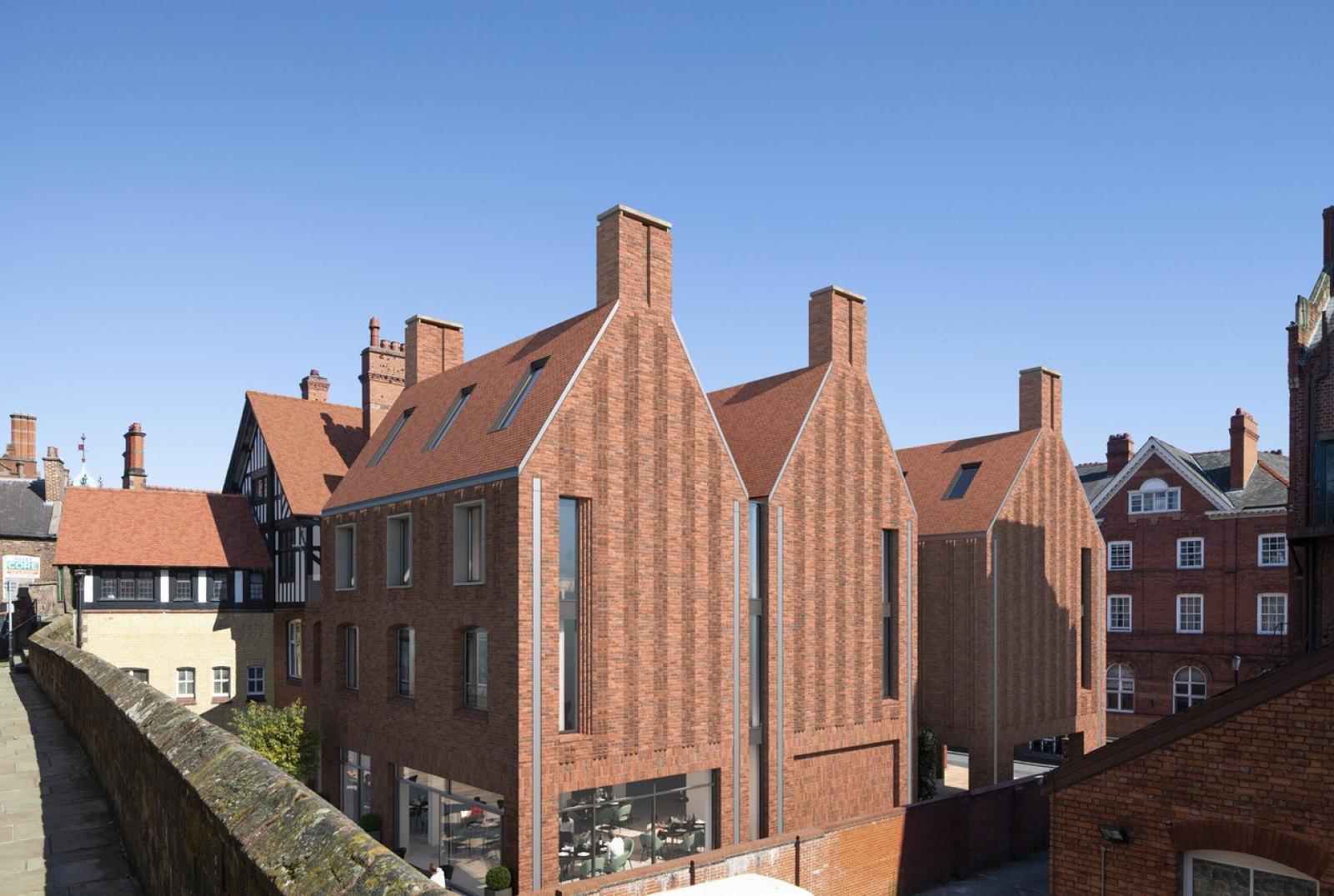 external image of a red brick hotel extension. The bricks have vertical bands which make the design striking and these lead up to its tall chimneys. The ground floor has a glazed cafe area with the first and second floors featuring six windows. The attic roof features three windows.