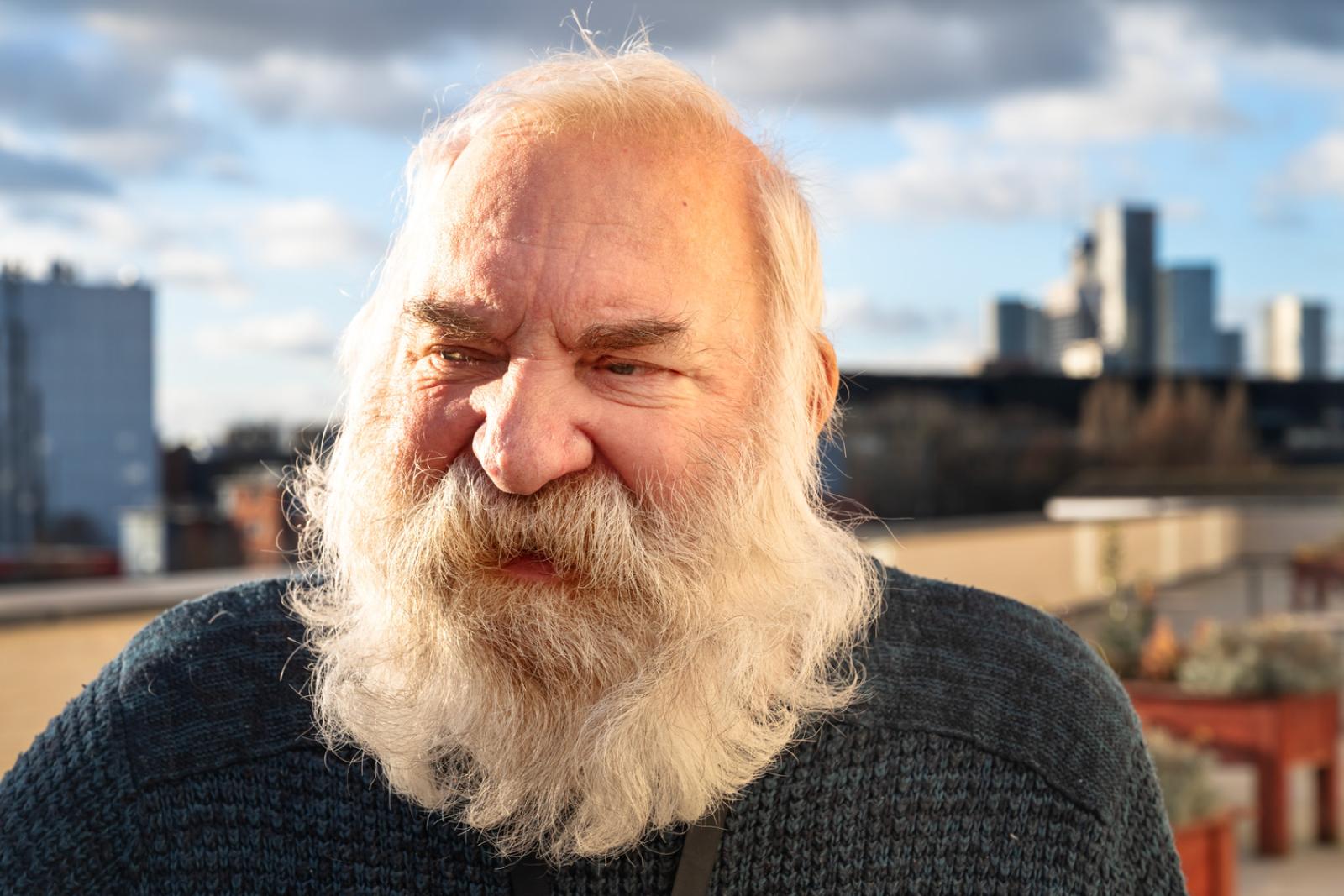 a man with long white beard and hair is in the foreground with the skyline of Manchester behind