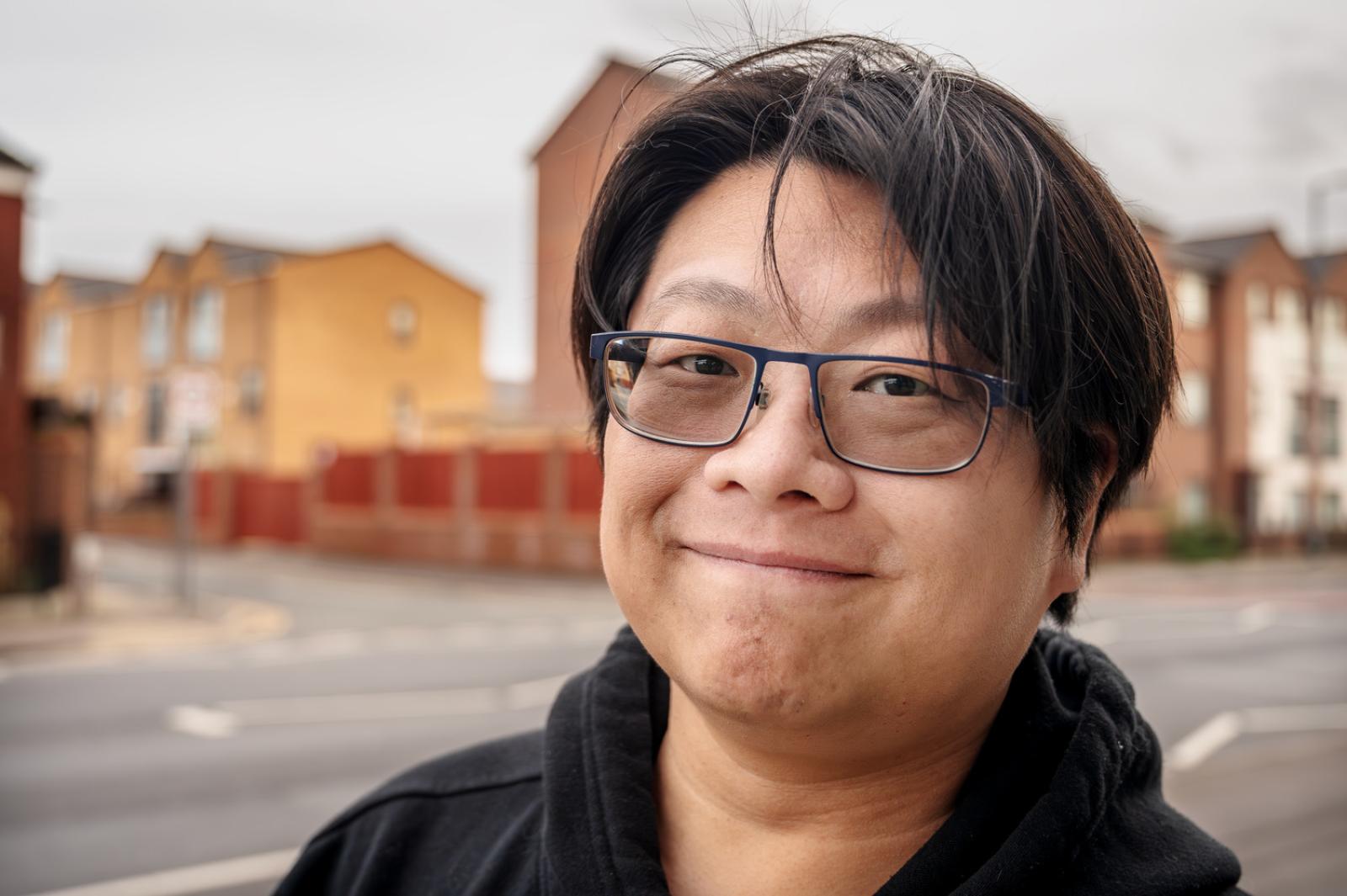 A portrait of a man wearing a black hooded top and glasses against the backdrop of Brunswick housing estate in Manchester