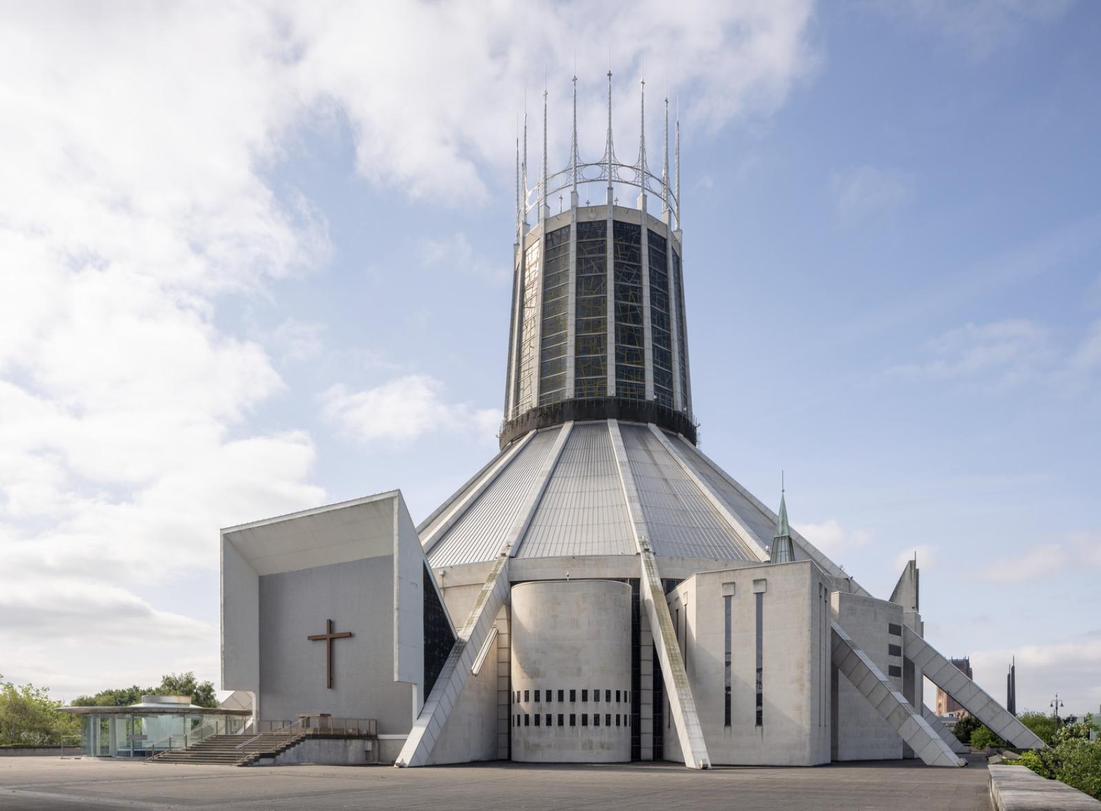 External photograph of a teepee shaped church made out of grey concrete. On the top half of the building there is a circular crown lantern made from coloured glass and finished with tall spires. 