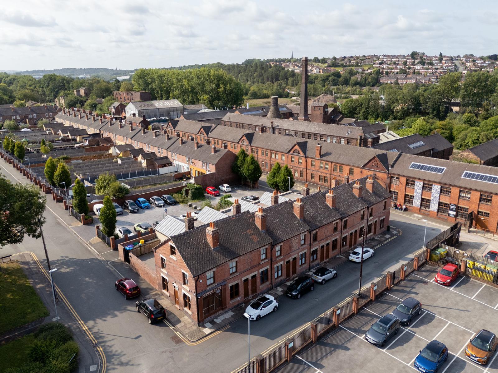 Aerial view of a historic red-brick industrial complex with rows of terraced houses in the foreground, a large mill building with a tall chimney in the center, and surrounding greenery and residential areas in the background. Several cars are parked along the streets and in a fenced parking lot.
