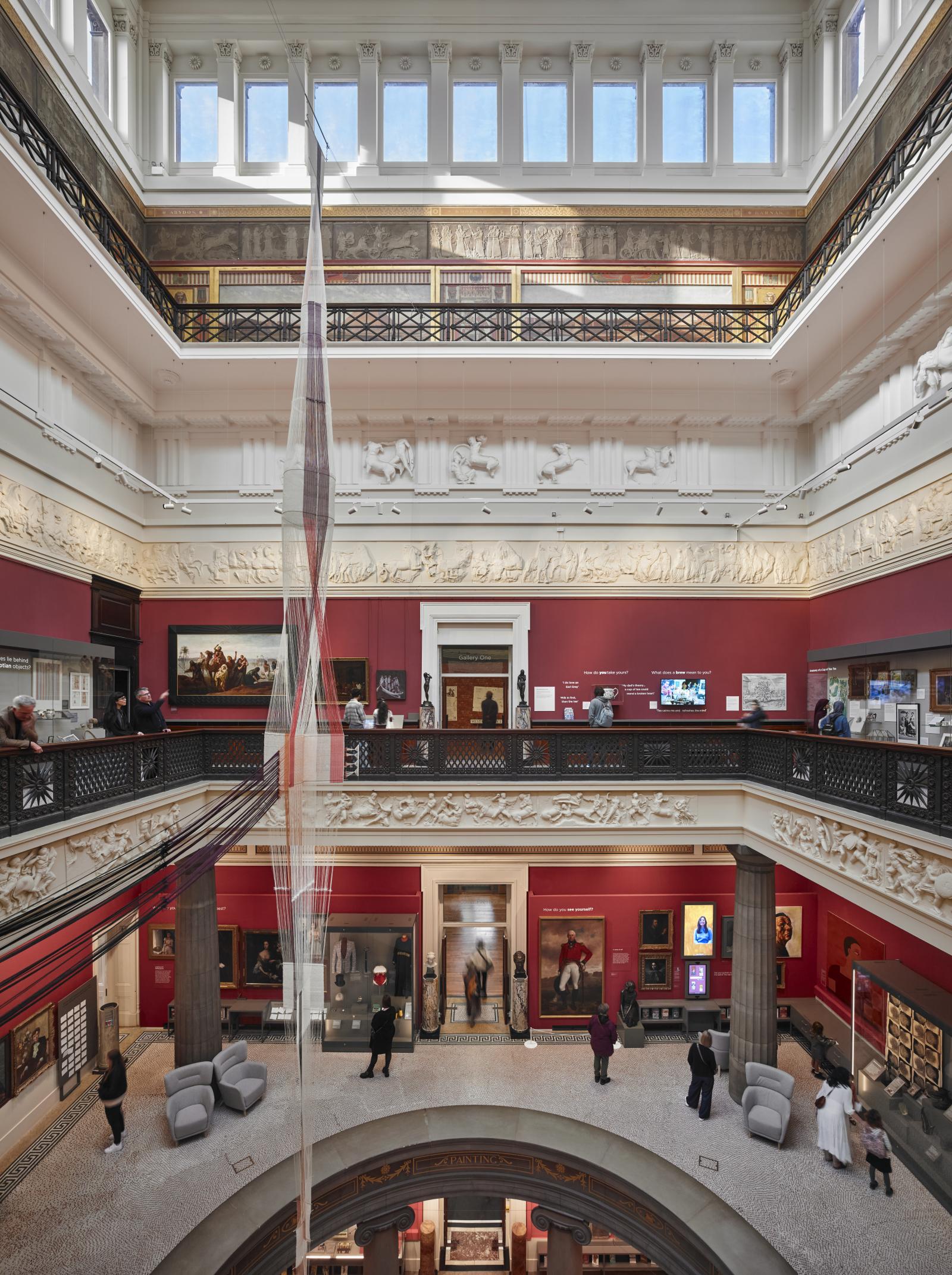 Interior view of a grand multi-level gallery with red walls, ornate white friezes, and classical columns. The space features paintings and artworks displayed on both floors, with visitors walking and observing exhibits. Natural light streams in from tall windows at the top level.