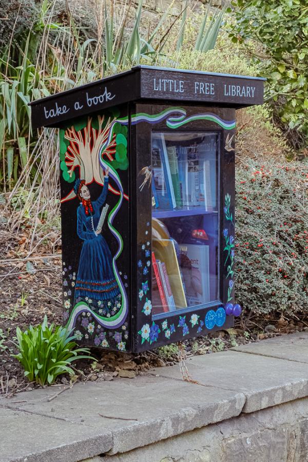 A small black cabinet with black folk art paintings used as a little free library on a morley street