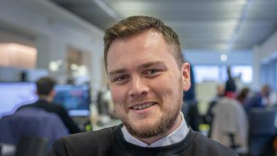 Headshot of Alex Scrimshaw. A man smiling, wearing a black jumper over a white shirt.