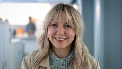 Headshot of Grace Avery. A woman smiling with short blonde hair, wearing a grey jumper and a cream jacket.