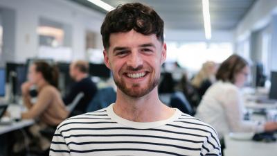 Headshot of Liam Briggs. A man smiling wearing a black and white striped top.