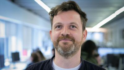 Headshot of Sebastian Drayson. A man smiling, wearing a black shirt over a white t-shirt.