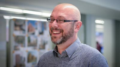 Headshot of Stephen Anderson. A smiling man, wearing glasses, a grey blue jumper and a checkered shirt.