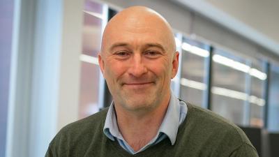 Headshot of David Parkinson. A man smiling, wearing a blue shirt and green jumper.