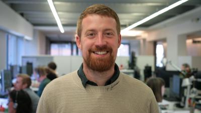 man in beige jumper smiling, standing in office