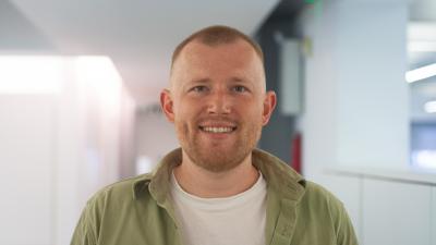 Headshot of Jake Ramsden. A man wearing a green shirt and white tee, smiling at the camera.