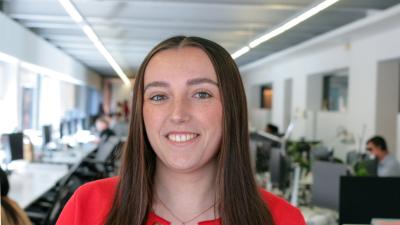 Headshot of Rebecca Williams, a girl with dark brown hair and a red cardigan, standing in an office.