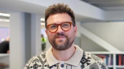 A portrait of a man in a patterned black and white cardigan with brown glasses