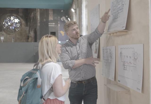 Internal photograph of a man showing a woman an architectural drawing on a noticeboard on a wall