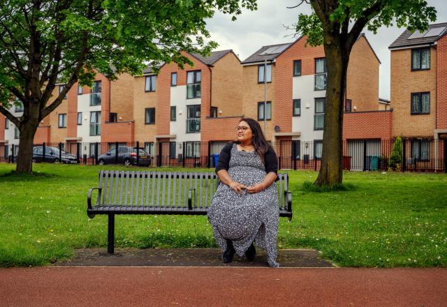 a lady sits on a bench in a park. A row of brick houses sit behind.