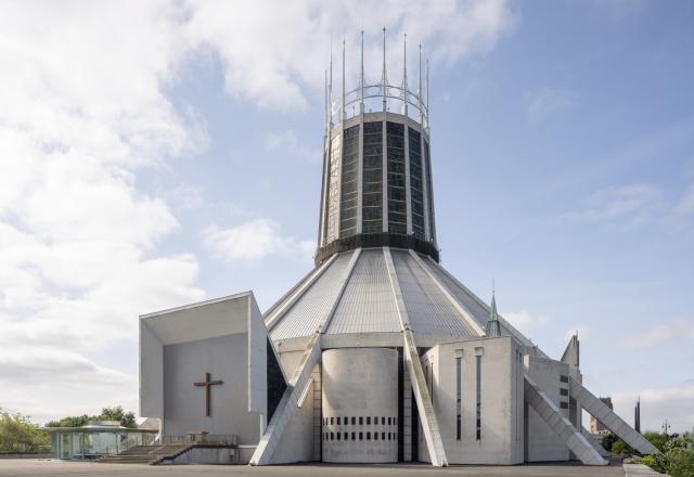 External photograph of a teepee shaped church made out of grey concrete. On the top half of the building there is a circular crown lantern made from coloured glass and finished with tall spires. 