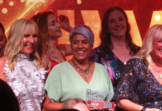 Image of women standing to receive applause at the end of an awards event. Chithra is centre in the image, smiling and holding her award. 