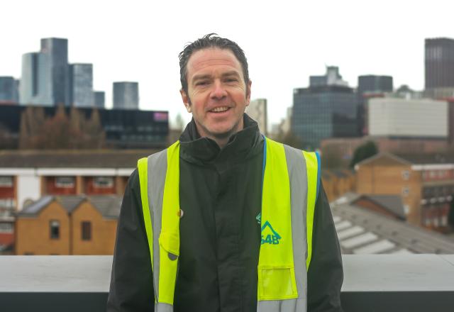 Photo of Ross Hemmings, a man with dark brown hair and a high visibility vest stood in front of the Manchester skyline