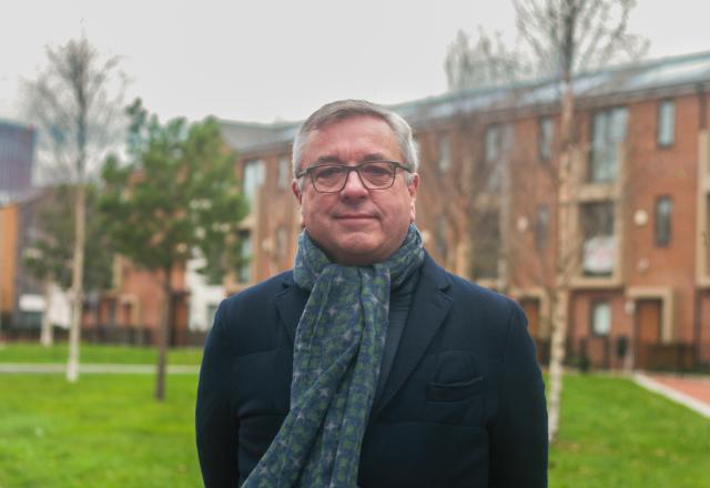 Headshot of Andy Avery, a man in a dark jacket and scarf, facing the camera in front of a green area and red brick houses.