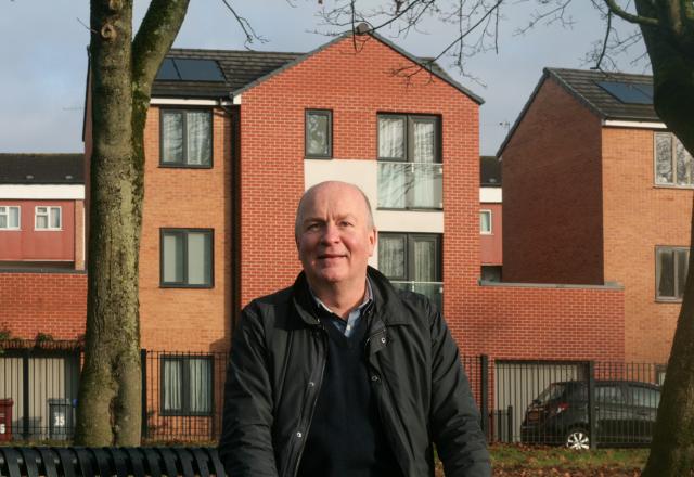 A man wearing a dark jacket sitting on a bench in front of a red brick building.