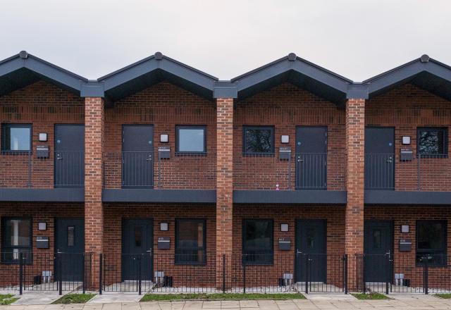 This image shows a modern row of red-brick terraced housing with gabled roofs, black-framed doors and windows, and small balconies on the upper floor. Each unit features a minimal front garden enclosed by a black metal fence, with a paved pedestrian area and street in the foreground. The design reflects contemporary urban housing, blending clean lines and traditional materials