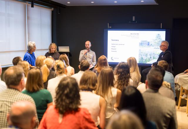 Audience of people facing four panellists and a presentation screen in a room.