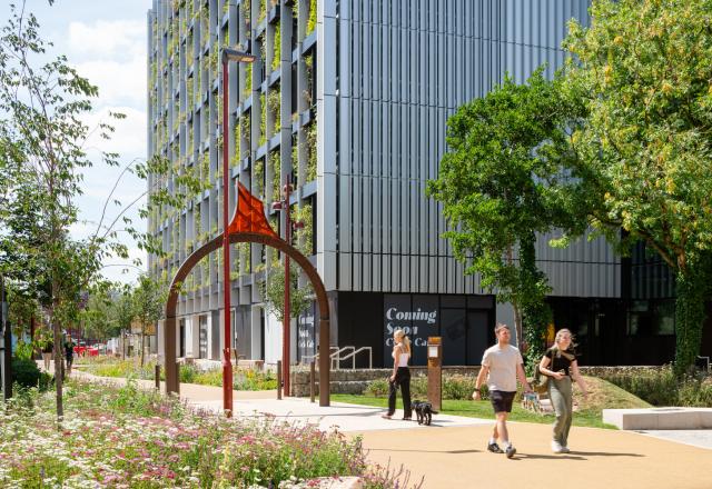 Modern building with integrated greenery, a flower-lined path, two people walking—one with a dog—and an archway sculpture; 'Coming Soon' sign visible.
