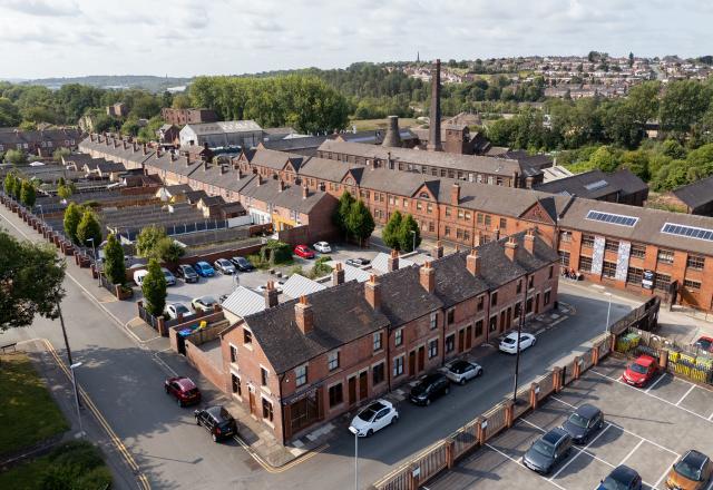 Aerial view of a historic red-brick industrial complex with rows of terraced houses in the foreground, a large mill building with a tall chimney in the center, and surrounding greenery and residential areas in the background. Several cars are parked along the streets and in a fenced parking lot.