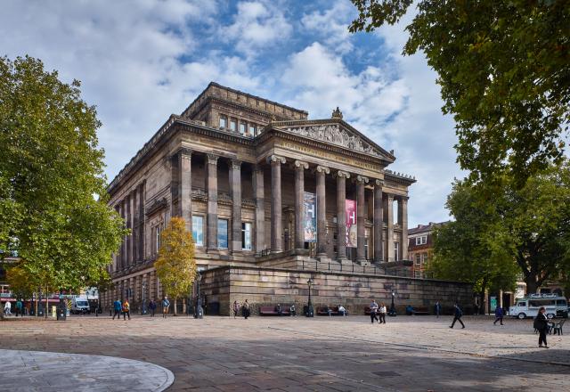 A neoclassical building with tall stone columns and ornate pediment, surrounded by trees and a paved square where people are walking.