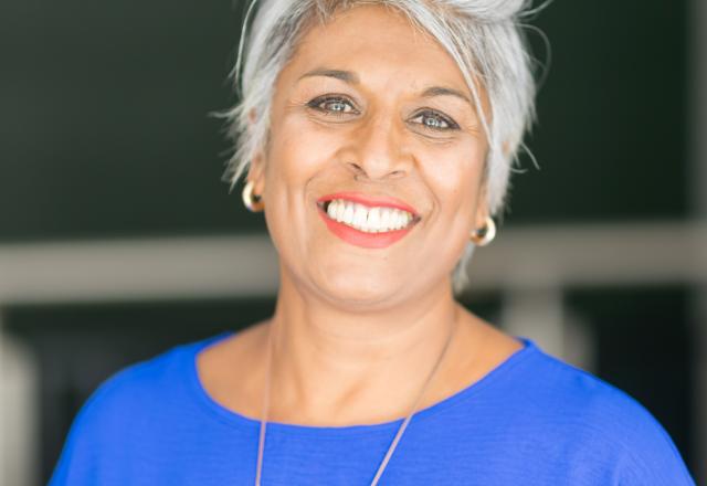Woman with brown skin wearing a blue top, short grey hair and smiling.