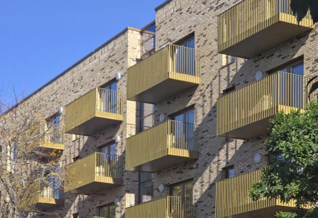 A residential apartment block over several floors with light brick and gold balconies, framed by trees.