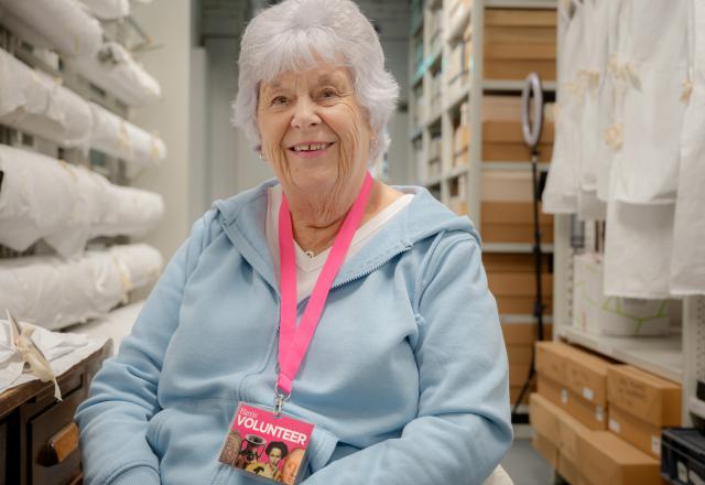 An elderly lady with grey hair, wearing a blue jumper and a pink lanyard stand down in a storage room.
