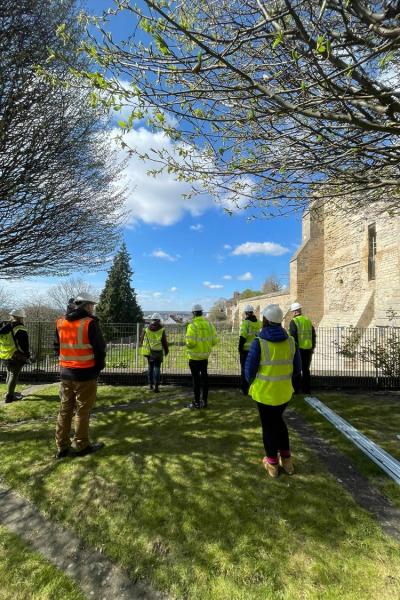 a group of people in hi vis stand on a lawn looking at a stone ruin 