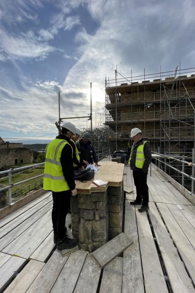 a small group of people in hi vis and hard hats stand on a scaffold looking at the top of a collumn