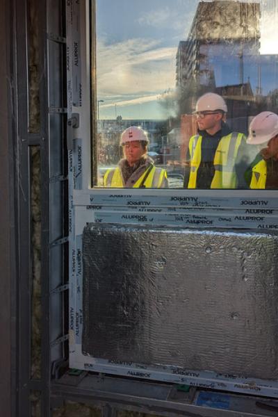 3 people in hi vis coats and hard hats are seen through the window of a sample unit on a building site