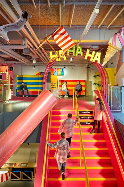 A scene inside the museum. An interior view of a brightly colored, modern space designed for children, featuring a bold pink staircase. Playful elements include a neon 'Ha Ha' sign, vibrant yellow and pink accents, and interactive zones with children actively using the space.