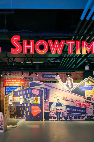 Front entrance to an entertainment venue or seaside shop featuring vibrant neon signage that reads 'It's Showtime!' The space is decorated with colorful murals, arcade-style designs, and a lively atmosphere, inviting visitors into the area