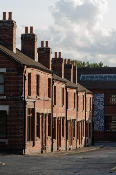 Row of terraced red-brick houses with pitched roofs and multiple chimneys, captured in angled sunlight. In the background, a larger industrial building with blue-and-white patterned panels and a skylight is partially visible.