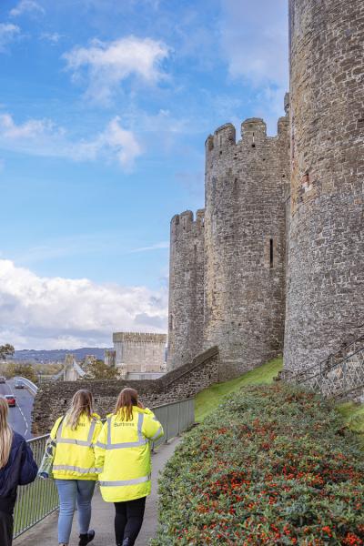 two ladies in hi vis jackets walk towards conwy castle on a sunny day