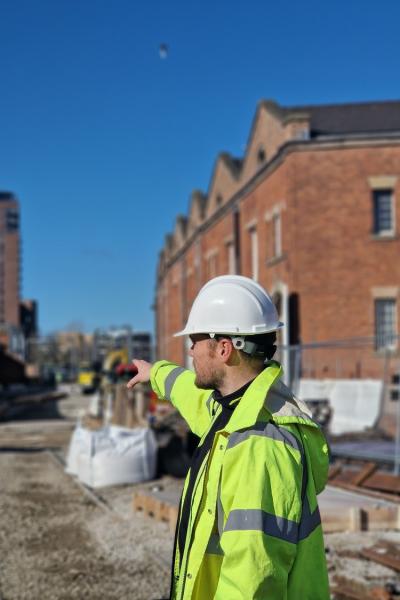 a man in a high vis jacket and hard hat is pointing towards some brick warehouse buildings