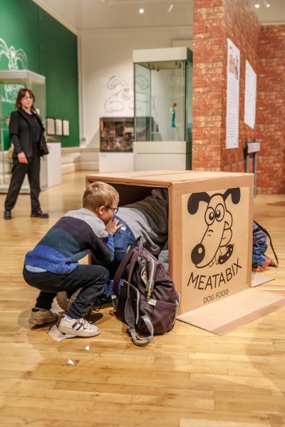 A child crouches near a large cardboard box labeled “Meatabix Dog Food” with a cartoon dog illustration. The box is part of an interactive exhibit in a gallery with wooden floors, display cases, and a brick-textured wall.